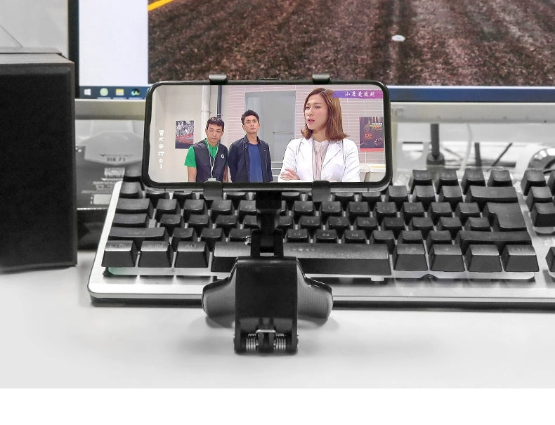 Phone mounted on a stand displaying a video call with three people, placed on a desk with a keyboard and computer monitor in the background.